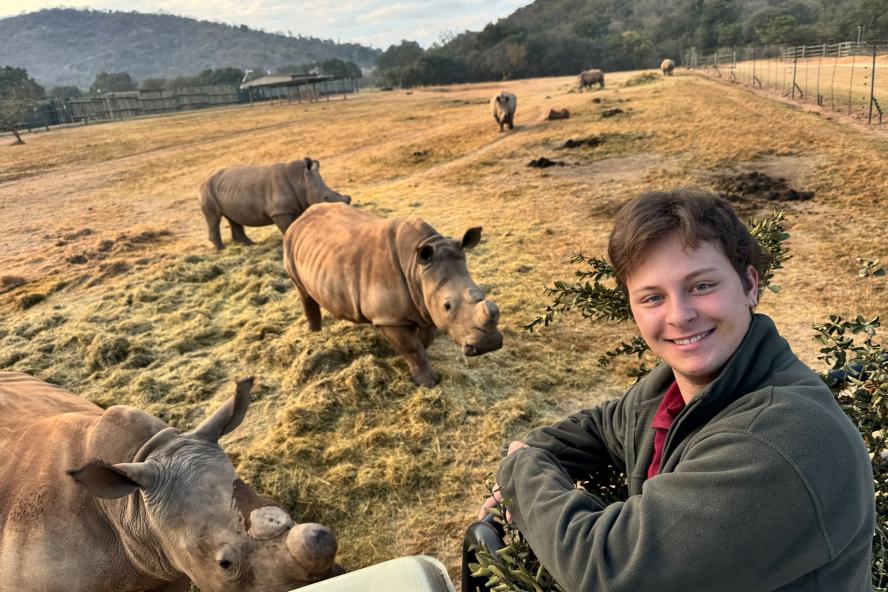 Kai Preminger in the foreground, smiling at the camera, with many rhinos in a field in the background.