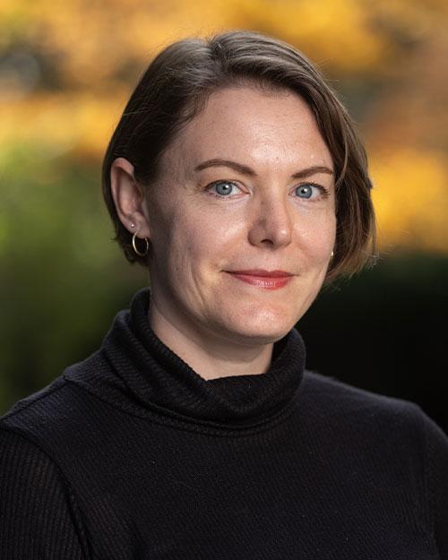 She is smiling at the camera, wearing a black turtleneck and gold earrings, with short brown hair and blue eyes.