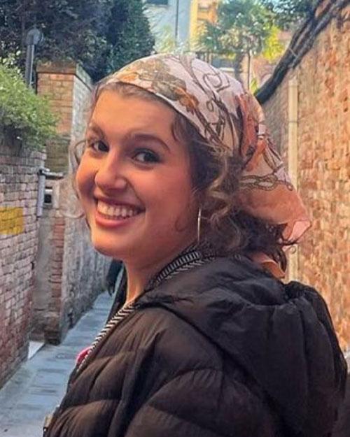 Photograph of Kathleen Landis. Kathleen is looking over her shoulder and smiling at the camera. She is wearing a patterned bandana, hoop earrings, and a black jacket.