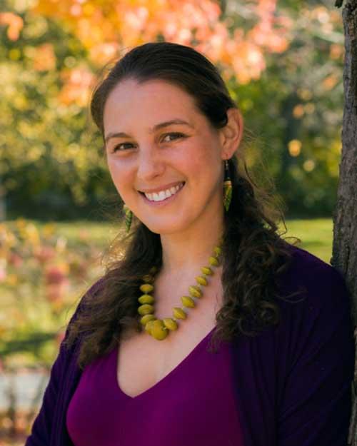 A woman is standing outside, leaning against a tree, and smiling at the camera