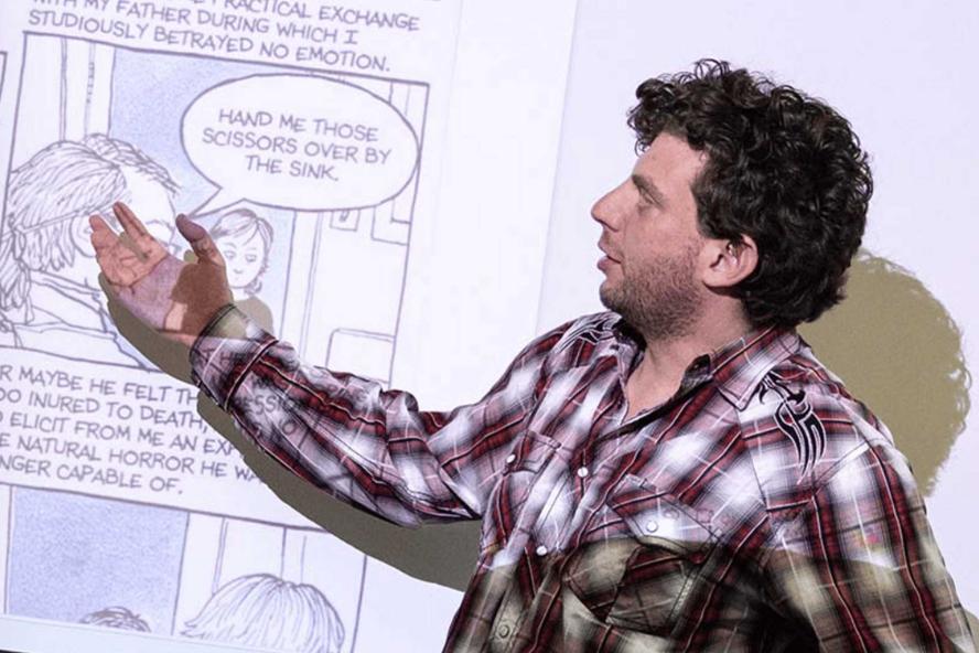 There is a man with curly brown hair wearing a flannel pointing to a comic strip that is being projected on to a wall