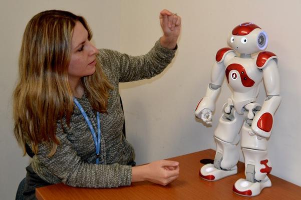 Woman with long brown hair and blue lanyard gestures toward a red and white robot on a table