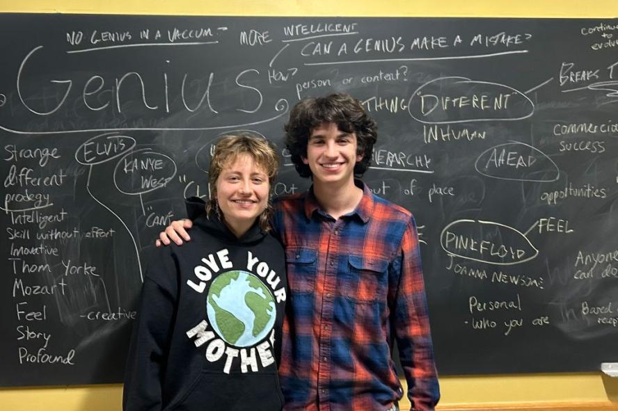 Two students smile at the camera in front of a chalkboard covered with writing