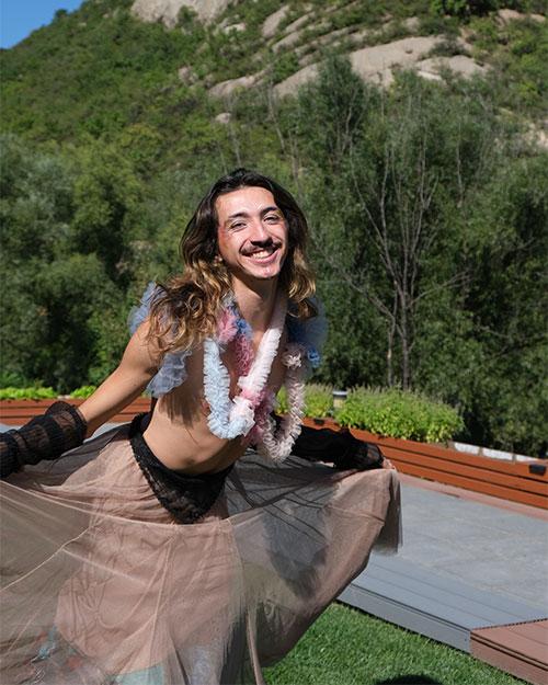 Person with long hair wearing a pink skirt and white leis stands outdoors, holding the skirt while posing in front of a mountain
