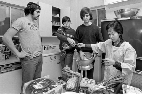 Four male students gathered around female chef at cooking table.