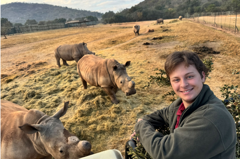 Kai Preminger in the foreground, smiling at the camera, with many rhinos in a field in the background.