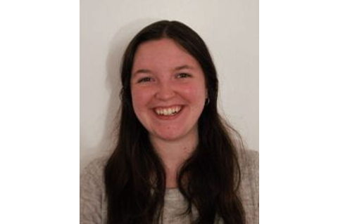 Brittany Velikaneye, with long, dark, brown hair, smiles widely at the camera, wearing a grey shirt and silver earrings