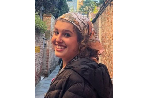 Photograph of Kathleen Landis. Kathleen is looking over her shoulder and smiling at the camera. She is wearing a patterned bandana, hoop earrings, and a black jacket.