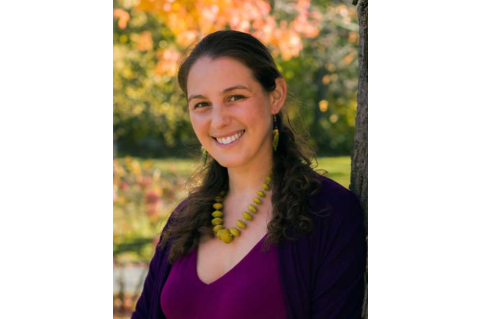 A woman is standing outside, leaning against a tree, and smiling at the camera
