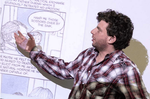There is a man with curly brown hair wearing a flannel pointing to a comic strip that is being projected on to a wall