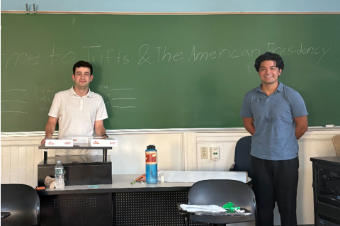 Two men in polo shirts smile at the camera while standing in front of a chalkboard