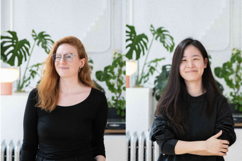 Two women in black shirts pose in a white room with plants; one has red hair and glasses, the other black hair